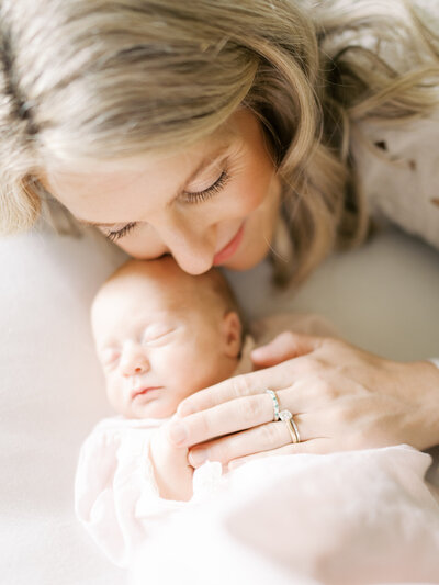 Blonde mother snuggles into newborn daughter wearing a pale pink heirloom gown while on a white backdrop.