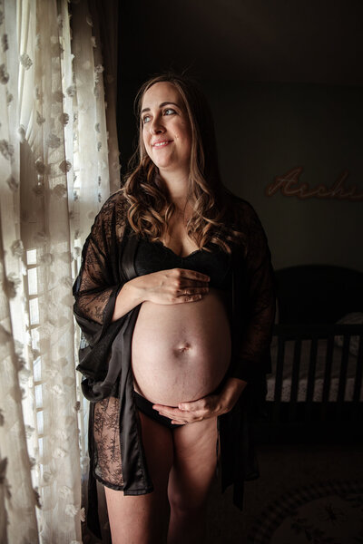 A pregnant woman stands in front of a window wearing a black lacy robe and black underwear and holds her belly as she looks out the window smiling