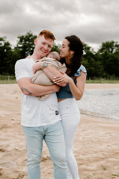 Dad in white shirt and jeans holding baby wrapped in tan blanket while mom is smiling and hugging dad wearing blue shirt and white jeans in Severna Park Maryland
