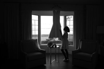 A silhouette of a Black bride gently adjusts her wedding dress, which hangs in front of a large bay window at Azaleana Manor in Orange Park, Florida. Captured in timeless black and white, the scene shows the bride in a robe and slippers, preparing for her wedding day as soft natural light floods the room. A bouquet and accessories rest on a nearby table. This quiet, emotional moment showcases the elegance of documentary wedding photography. Photographed by Phavy Photography, a St. Augustine wedding photographer known for editorial storytelling and heartfelt bridal prep imagery.