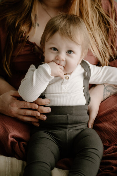 Baby boy in brown overalls and white top sitting on moms lap in studio in Severn Maryland.