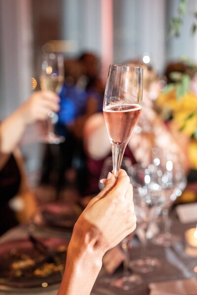 A hand raising a glass of sparkling pink champagne in a toast, surrounded by blurred people and a table set with plates and other glasses, suggesting a celebratory or festive atmosphere.