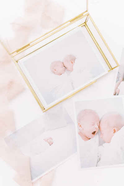 Image of photographic prints of newborn twins laying scattered on white backdrop and in gold and glass box with pink ribbon image by Portland Newborn Photographer Emilie Phillipson Photography.