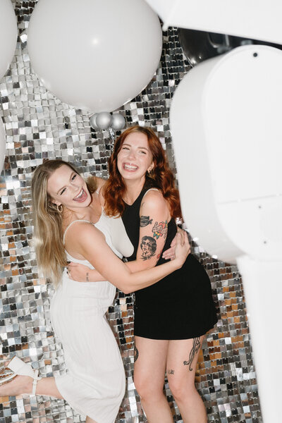 Two women laughing and hugging in Always Present photo booth against a modern disco backdrop.
