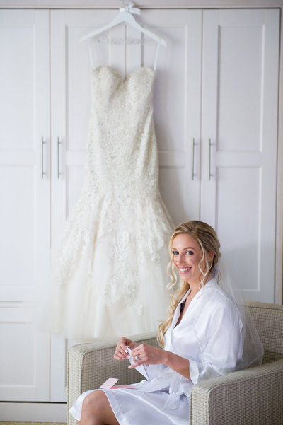 Bride sitting in a robe in front of a white wall of cabinets with her wedding gown hanging as she smiles after reading a note from her groom. 