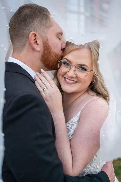 Groom kissing bride’s forehead while she smiles at camera