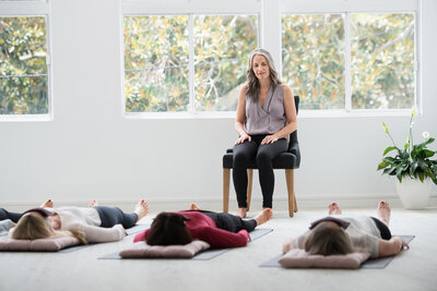 Marcia in a chair presenting a Yoga of Stillness session with participants laying down