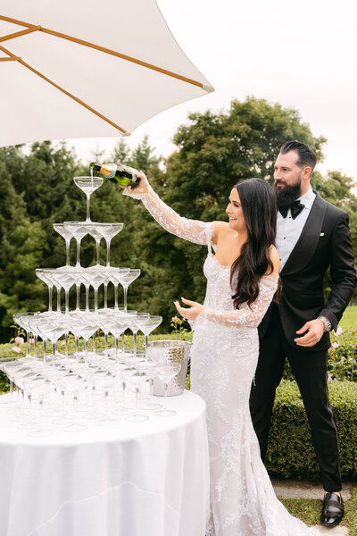 A bride pours a bottle of champagne in the top glass of a champagne tower at an outdoor venue