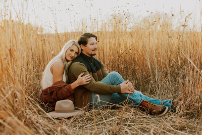 couple sitting in tall grass with hat during fall engagement session in lincoln nebraska, photo by kaitlyn neeley, wedding photographer in omaha