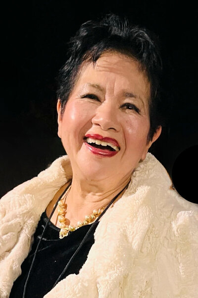 Author, speaker, and life coach Nita Tin at an event in Trilith, Georgia, wearing a black dress with a light-colored coat and a gold and pearl necklace, smiling warmly against a black background.