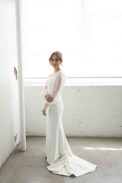 Person standing in white wedding dress looks back towards camera and smiles