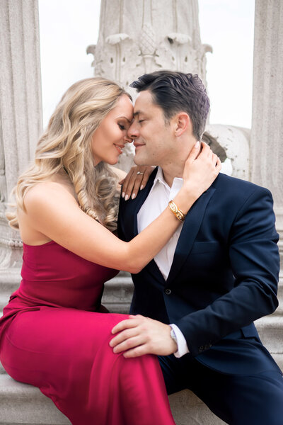 A couple in elegant attire shares a tender moment. The woman, in a red dress, gently holds the mans face, who is in a dark suit. They are seated in front of a decorative stone wall, gazing into each others eyes.
