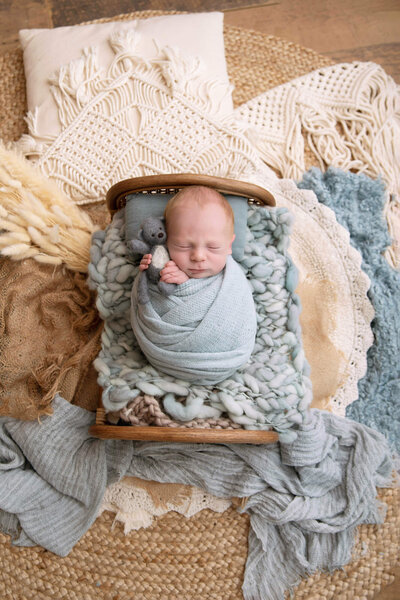 newborn boy wrapped in blue on a tiny bed holding a blue teddybear in a boho setup for his newborn photography session in Burlington, Ontario.