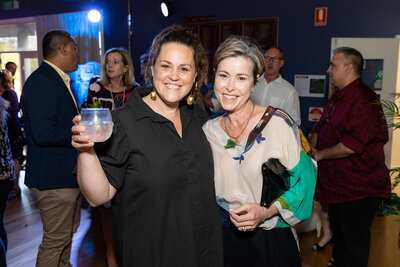Two ladies at a Sydney Event smiling towards the camera. The background appears to have a party going on and is dark. One of the ladies hold a drink in her hand.