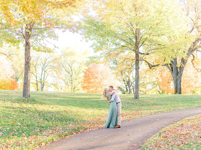 Future bride in a sage green floor length dress passionately kisses future groom in a white buttonup shirt and tan slacks with fall colors behind them