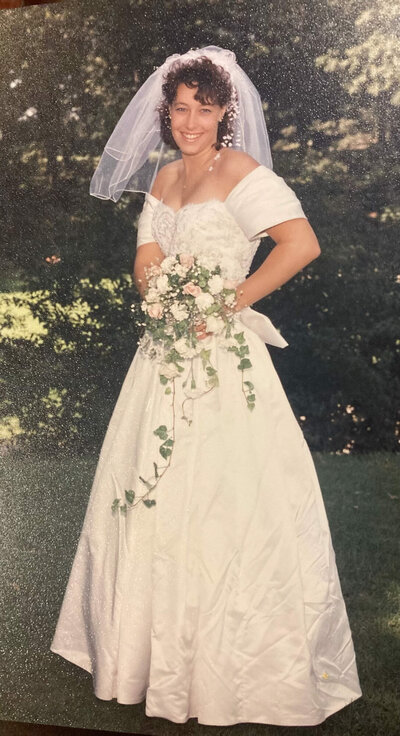vintage photo of a bride with curly hair, smiling and holding a bouquet, wearing a white dress with off-shoulder sleeves and a long veil, set against a lush green background