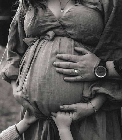 Cherish the quiet anticipation of new life with a poignant maternity session by Everest Portraits, highlighting the loving touch of family hands on a pregnant belly, presented in classic monochrome for timeless appeal.