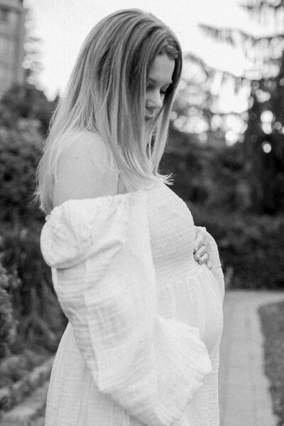 A pregnant woman in a flowing white dress cradles her belly while gazing downward, set against an outdoor backdrop. Photographed in Toronto, Ontario.