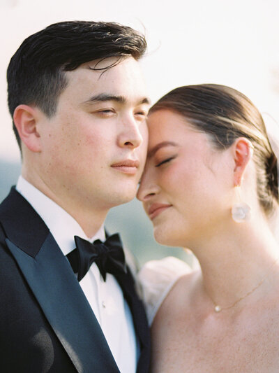 A close up portrait of a Korean-American bride and groom leaning their heads towards each other while the groom looks out over the horizon