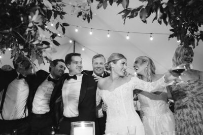 Black and white image of bride and groom with their arms around wedding party and guests under string lights