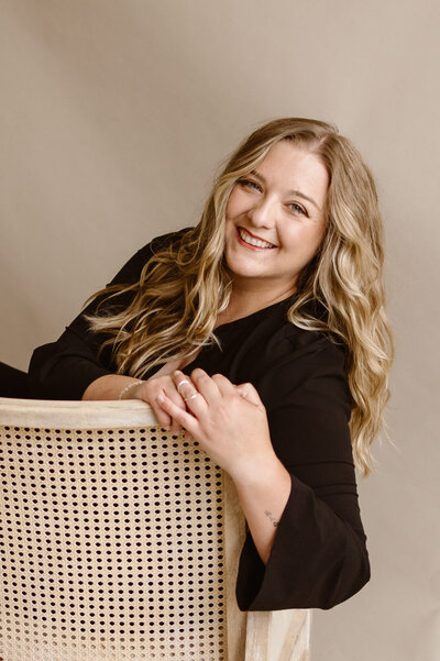 Cassandra, a Denver photographer, smiles while leaning over the back of a woven cane chair, wearing a black top against a neutral studio backdrop.