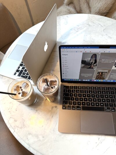 A neatly stacked pile of light-colored books, primarily white and beige, sits on a surface. A lit black jar candle rests on top of the books. Behind the stack, sheer net curtains add a soft, diffused light to the scene.