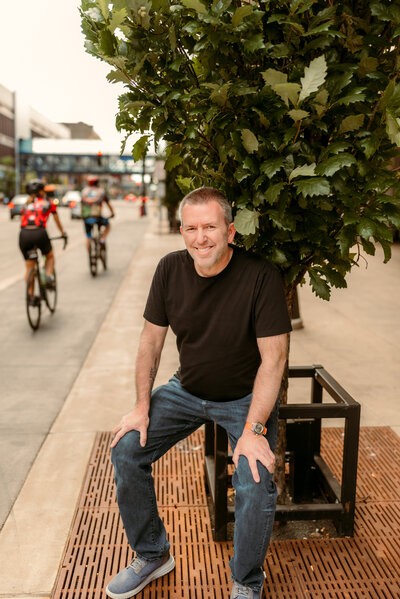 Casual outdoor dating photo of Kerry in downtown Minneapolis, sitting relaxed in a black shirt with city life in the background.