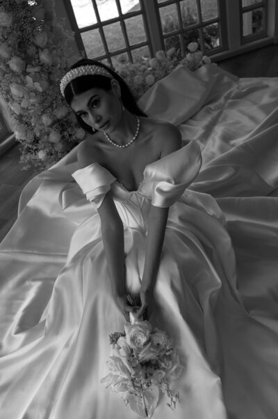 A bride in a luxurious satin gown with puffed sleeves sits gracefully, holding a bouquet of white flowers. Bright windows illuminate the scene.