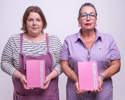A picture of two humans on a lilac background. Lucy on the left is wearing dungarees and a striped t-shirt and has shoulder length brown hair. Pippa on the right is wearing a lilac jumpsuit, has a close cropped salt and pepper hair. Both have deadpan expression and are holding pink notebooks with the words "disobedient fuckery".