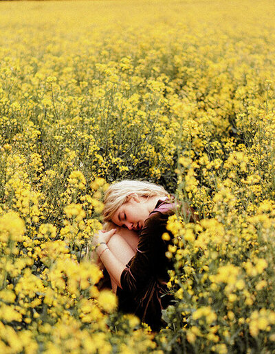 Jeune femme immergée dans un champ de fleurs jaunes, vêtue d'une robe en velours marron, genoux contre sa poitrine.