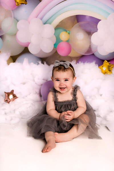 Portrait Photographers CT photograph joyful baby girl in gray tulle dress against pastel rainbow balloon backdrop with fluffy clouds