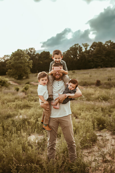 Dad in a henley shirt and khaki pants holds his three sons in his arms and on his shoulders while in a field at sunset in cowboy boots