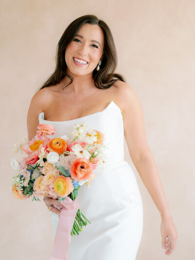 Portrait of bride in a strapless dress holding a colorful bouquet in front of a light pink wall