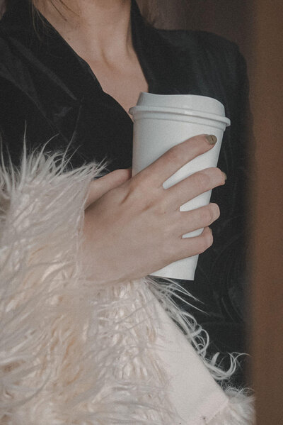 A brand strategist in a black top and a light-colored furry coat holds a white disposable coffee cup with a lid—perfectly embodying the creative vibe of Sweet Pea Ink Creative.