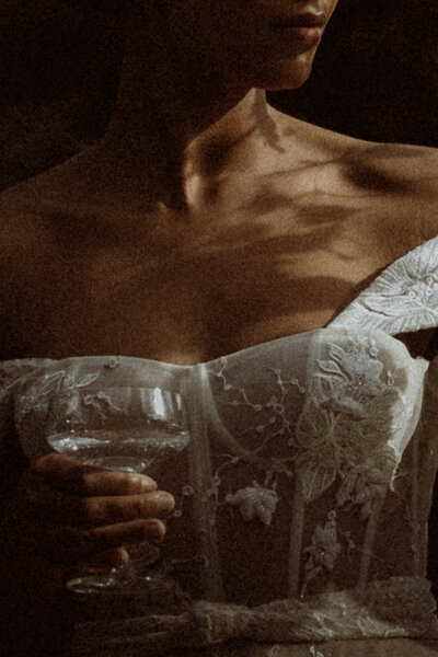 Detail shot of a bride's lips down to her waist. There's shadows of her bouquet on her chest and she's holding her champagne flute while her head is faced to the side.