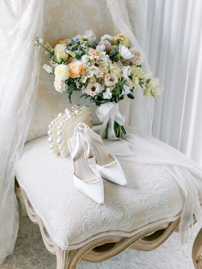 Elegant bridal bouquet of soft peach and ivory blooms with Bella Belle heels and a pearl clutch styled on a vintage cream chair.