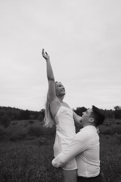 Groom lifting bride during playful engagement session at Sterling Nature Center in Central New York, surrounded by open fields and natural landscape