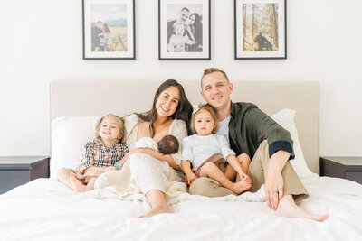 a family with two toddler girls and a newborn sit on a bed and smile at the camera of their Austin newborn photographer.