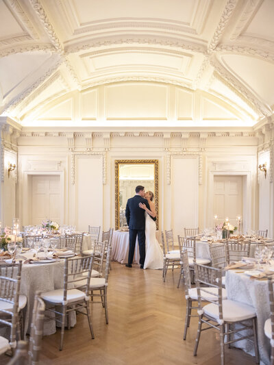 A couple embraces in a beautifully ornate ballroom with a high ceiling and decorative moldings. The room is set up for an event with round tables covered in white linens and surrounded by elegant chairs. A large mirror adorns the wall behind them.