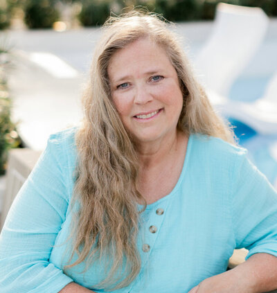 A woman with long wavy hair wearing a light blue top smiles while sitting outdoors near a pool or patio area.