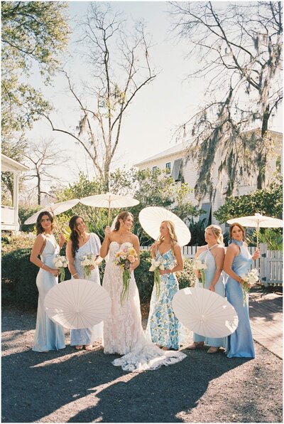 A bride laughs with her bridesmaids in blue dresses as they all hold white umbrellas in a garden captured by a Wilmington wedding photographer