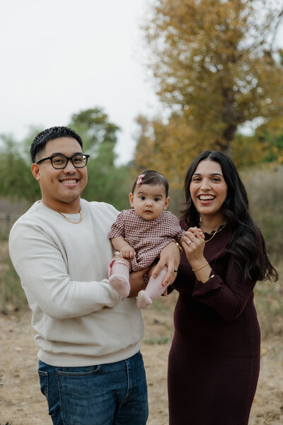 Smiling parents holding their baby girl during a cozy fall family session in a wooded area.