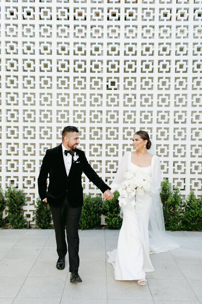 Husband and wife hold hands and stare lovingly at each other in  front of the breezeblock wall