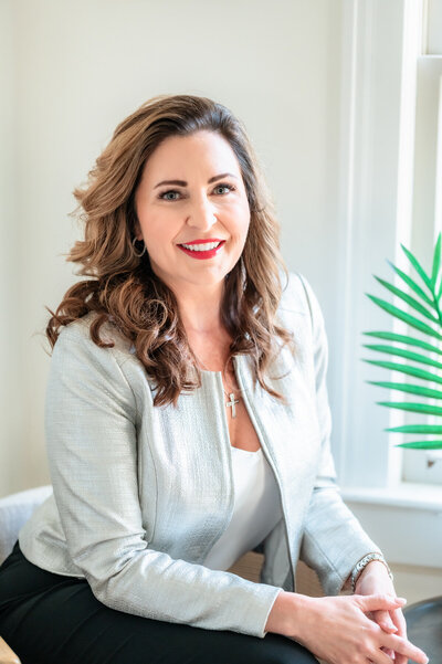 A woman with long, wavy brown hair wearing a light gray blazer and white top, seated near a window with a plant visible in the background, smiles at the camera.