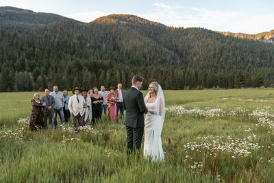 A couple exchanges vows in a wildflower meadow surrounded by friends and family with forested mountains in the background, captured by Sydney Breann Photography during their intimate Montana wedding.