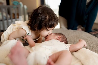 a mother holding newborn baby laughs with mouth agape looking at her partner.  a crocheted item is hanging behind her.