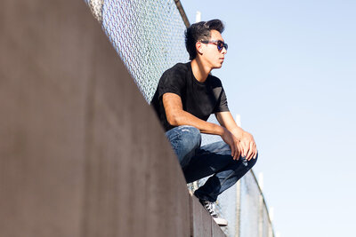 Boy wearing a pair of sunglasses squatting next to a fence.