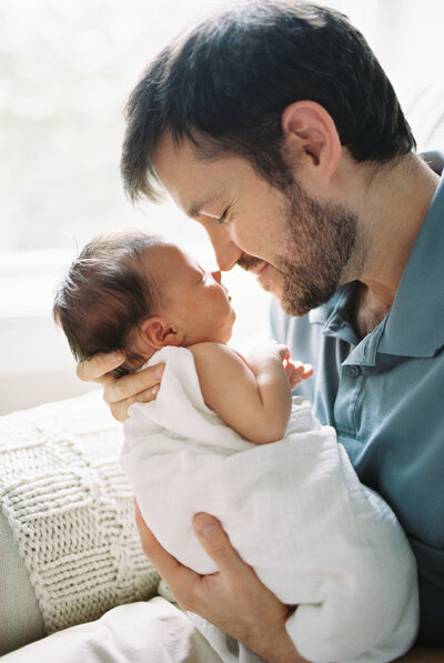 A father holding his swaddled newborn baby and touching his nose to the baby's nose during a photo session with Boston family photographer Corinne Isabelle