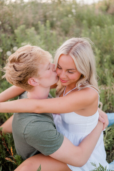 A couple poses for the camera in an open field with woman hugging man as he kisses her cheek.