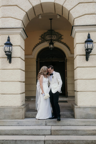 Die Braut flüstert ihrem Bräutigam vertraut etwas ins Ohr. Dabei steht das Paar entspannt unter einem Torbogen vor einem historischen Gebäude.